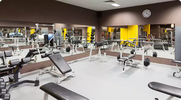Empty gym interior with benches, weight racks and dumbbells; mirrored walls reflect a yellow accent wall and a wall clock.