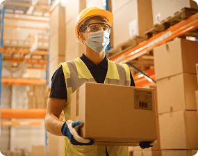 Worker in a safety helmet and mask carrying a cardboard box in a warehouse, symbolizing safe glass packaging and fast delivery.