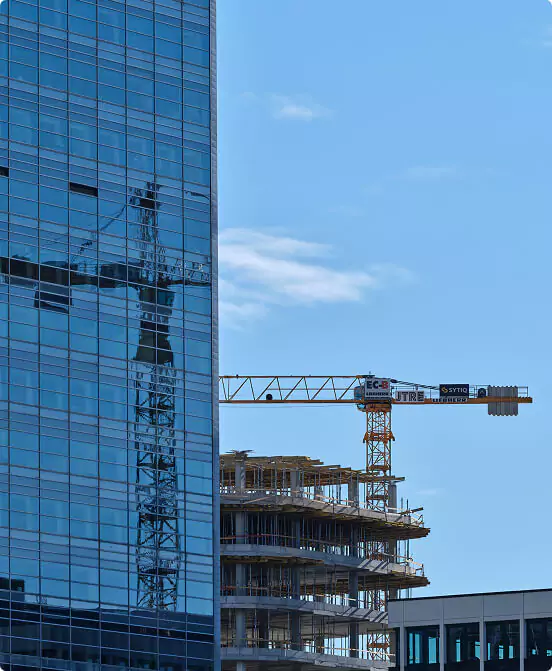 Glass high-rise reflecting crane beside construction site under clear blue sky.