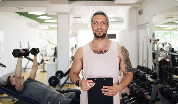 A smiling man with a clipboard stands in a gym behind him, another person lifts dumbbells on a bench surrounded by weights.